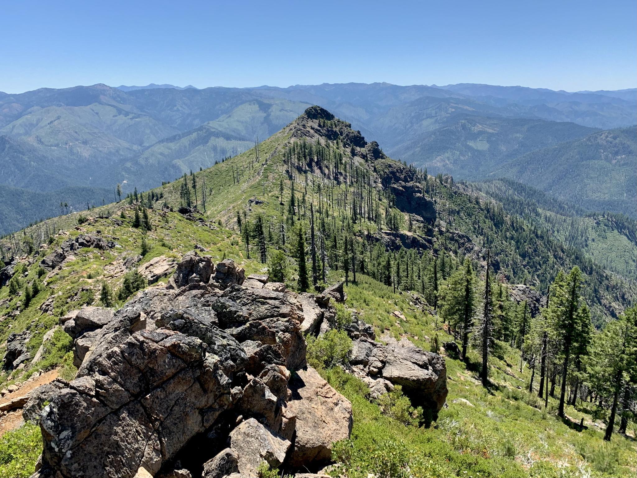 Sunny view of a rocky, forested mountain ridge and distant peaks under a clear sky.
