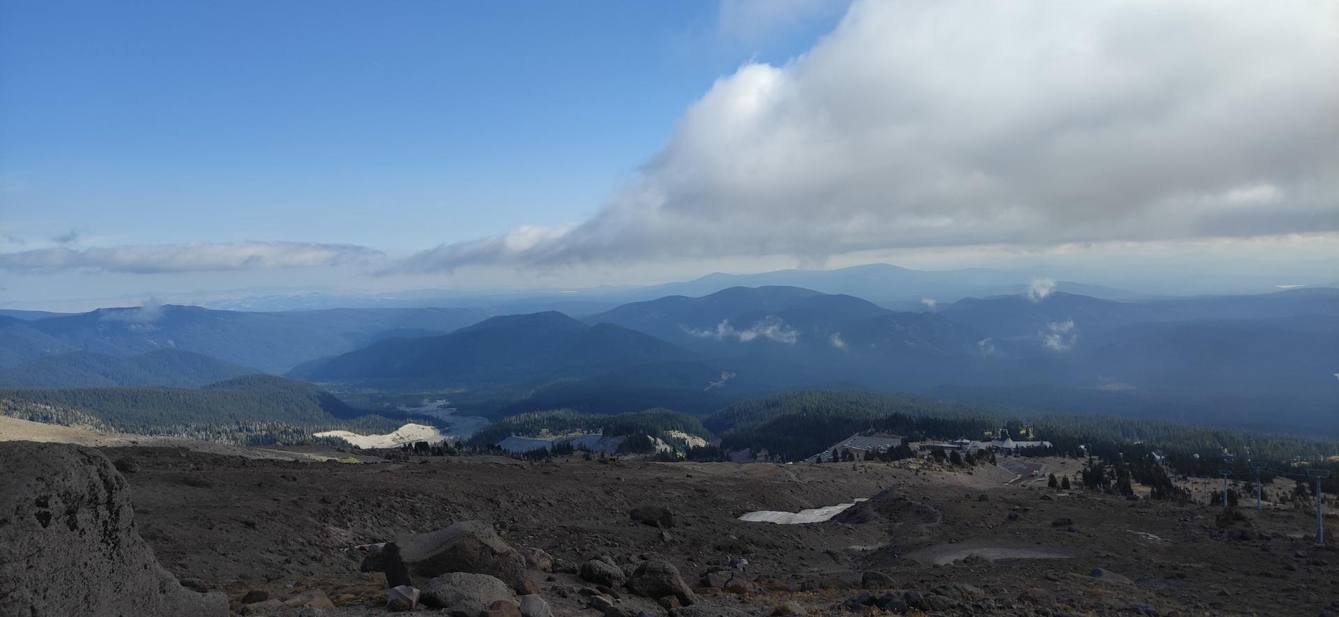 Vast mountain landscape with distant lake under a partly cloudy blue sky.