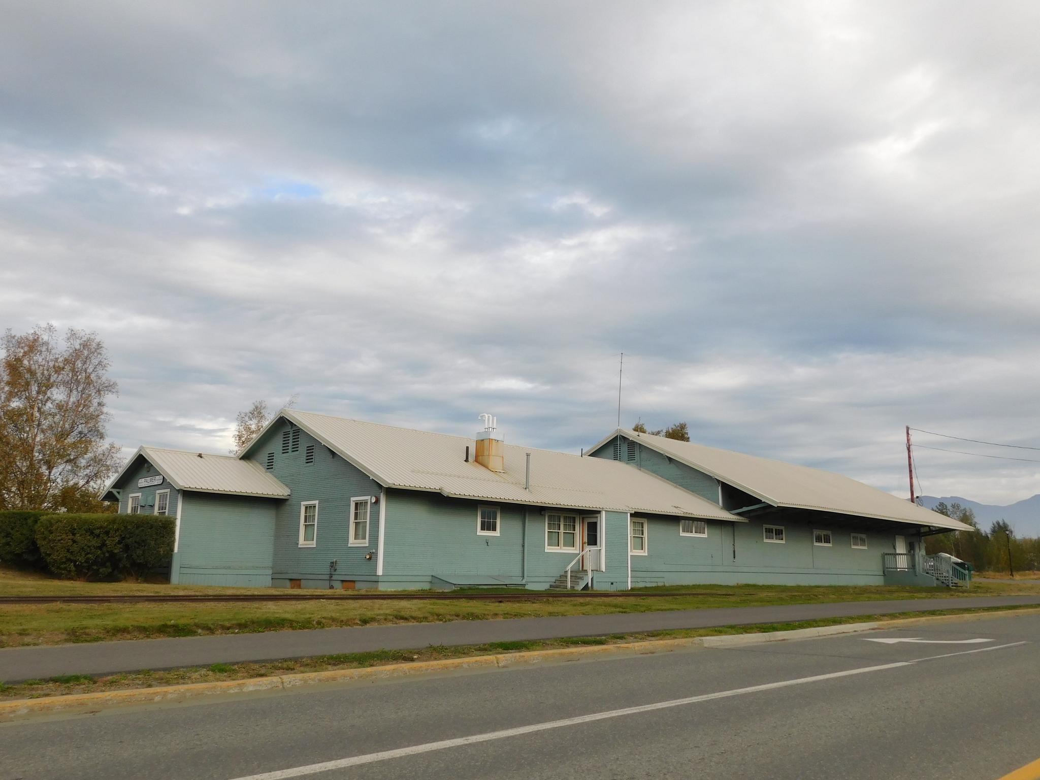Long light blue building with light roof under an overcast sky, next to a road.