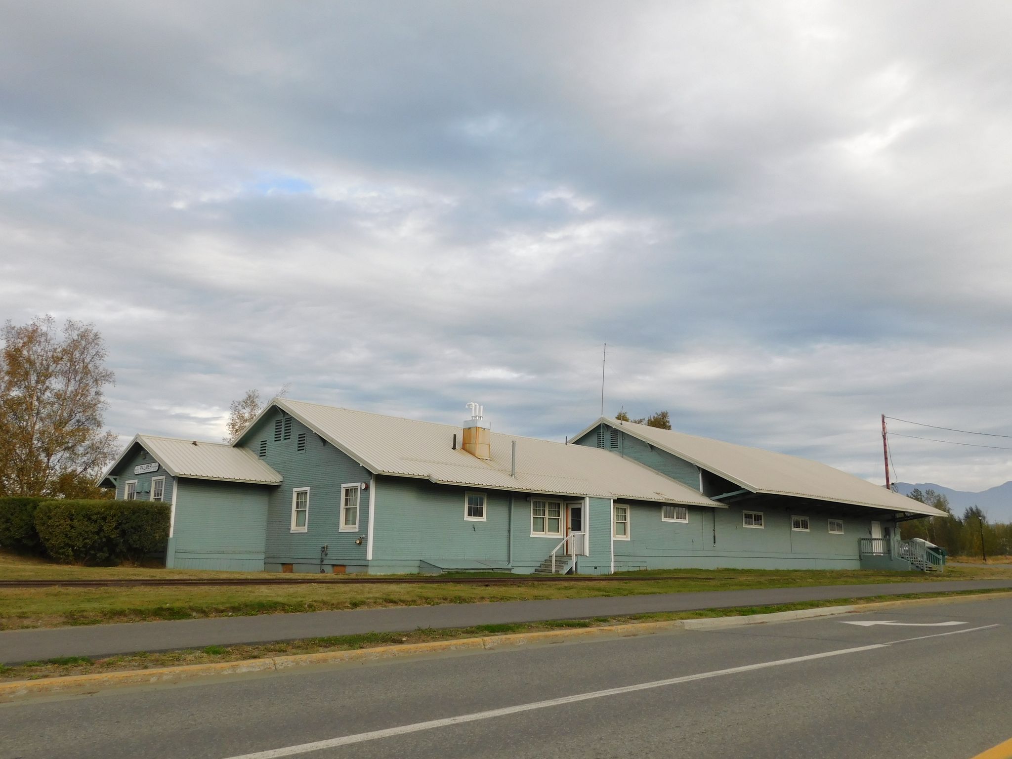 Long light blue building with light roof under an overcast sky, next to a road. - Train Depot - PHS Trails Loop