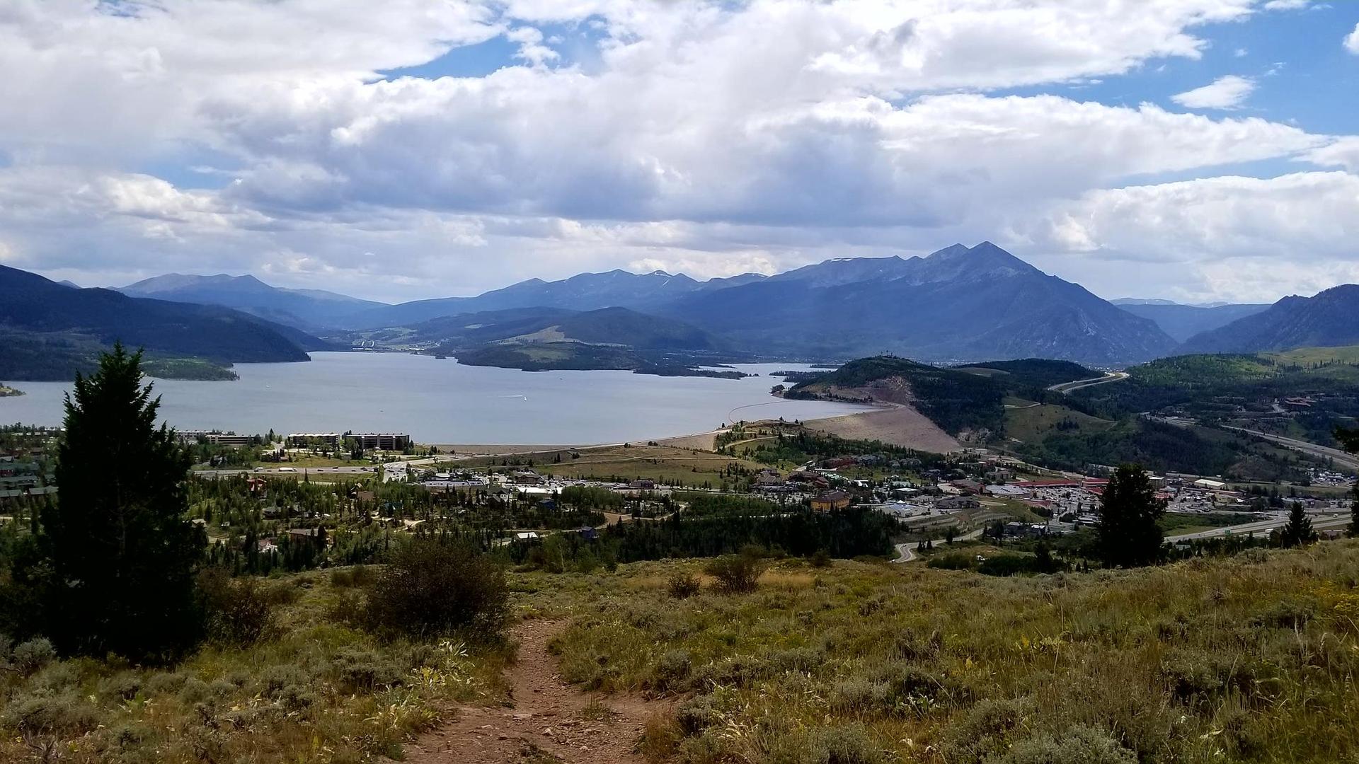 A dirt path leads to a scenic lake, mountains, and a distant town under clouds.