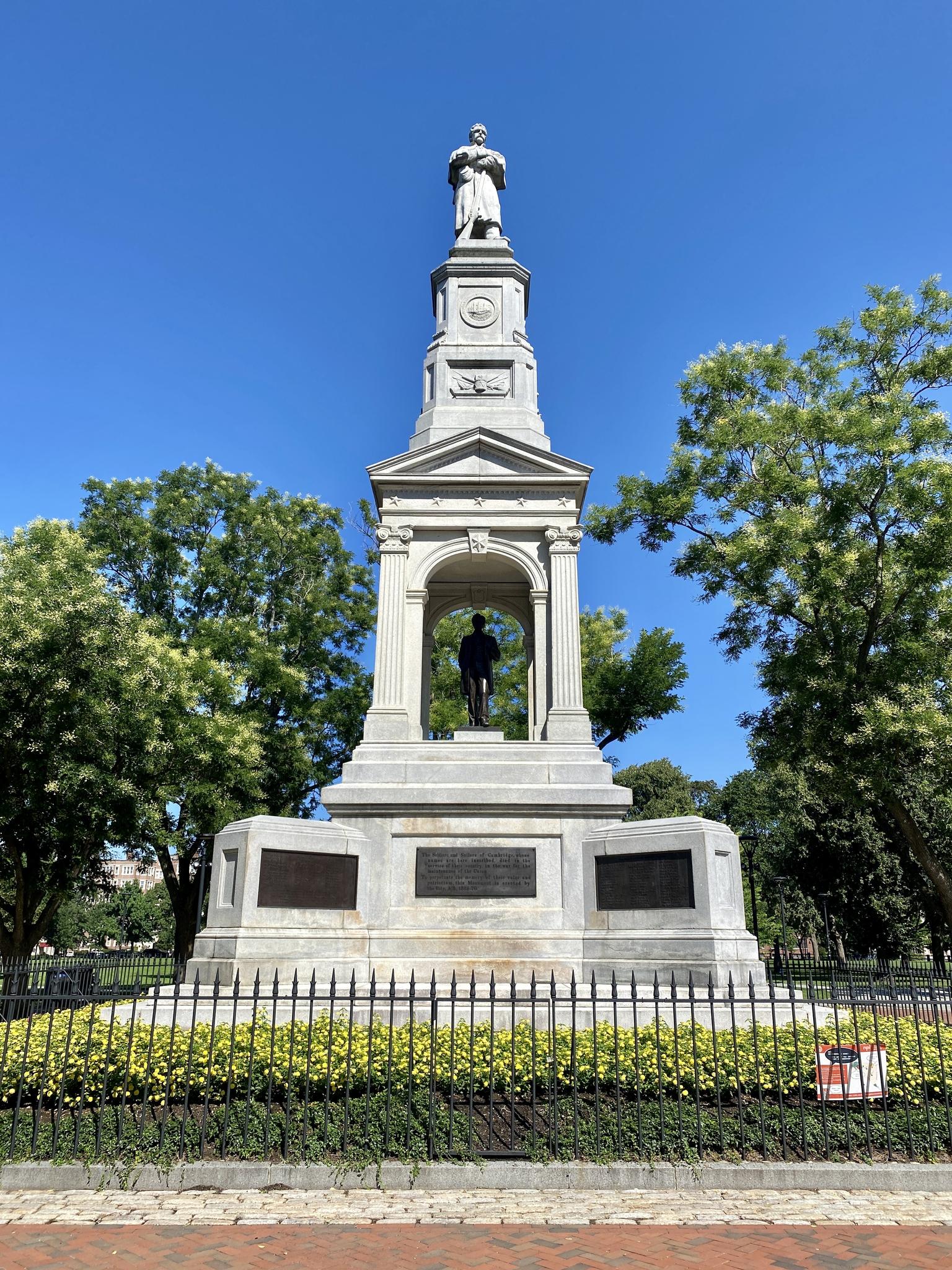 Tall white stone monument with statues, surrounded by trees under a clear blue sky.