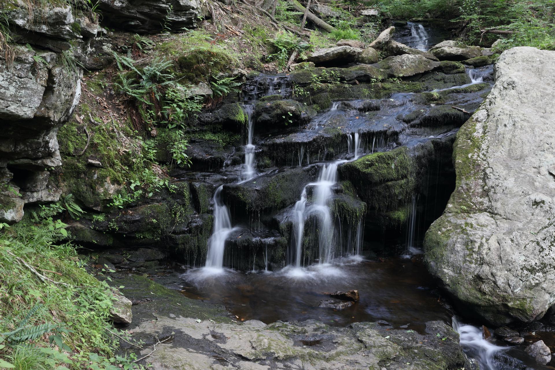 Multi-tiered waterfall cascades over mossy rocks in a shaded forest.