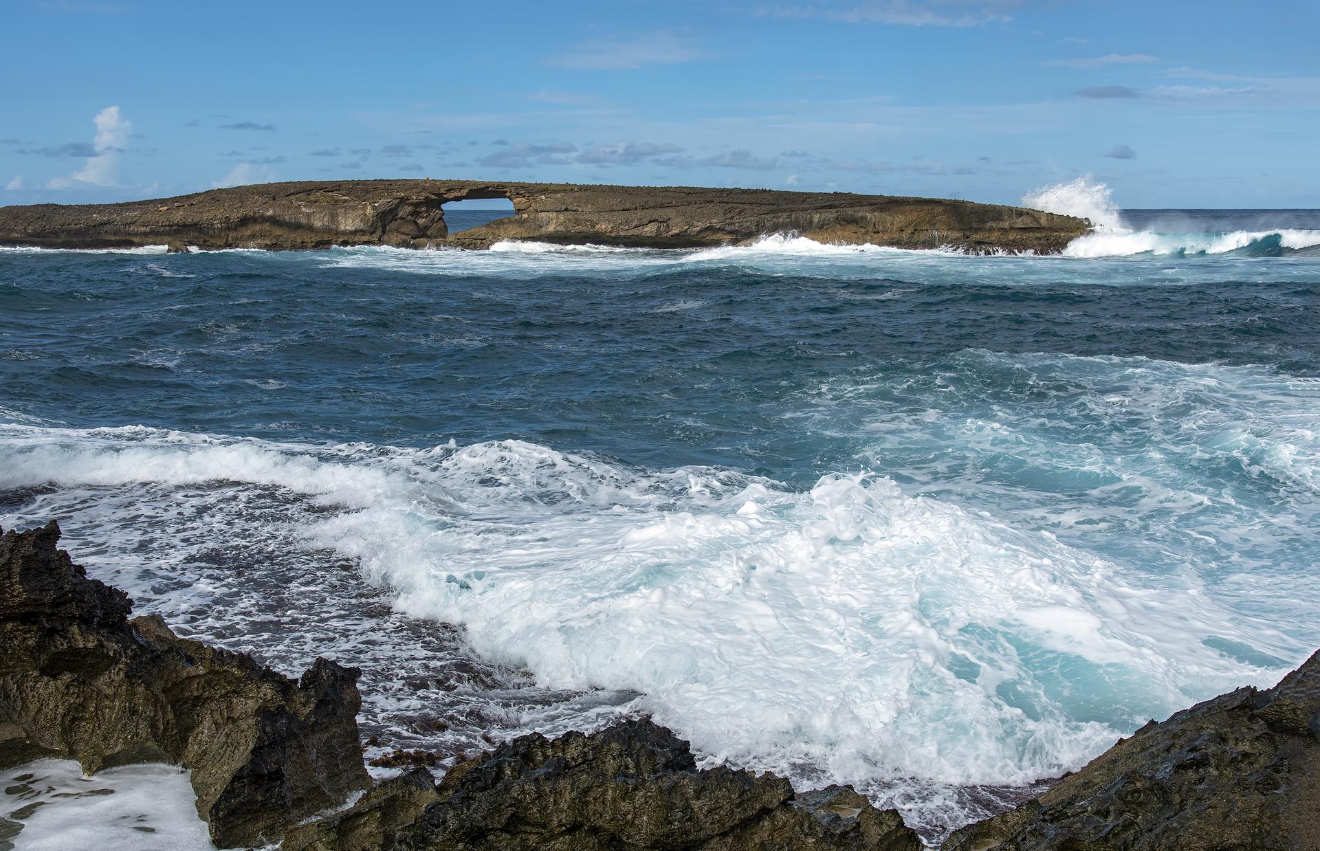 Rough ocean waves crash on a rocky shore with a natural arch in the distance.