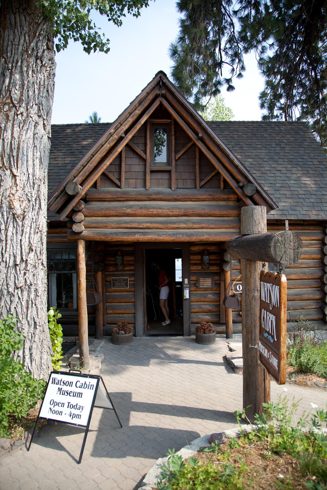 Rustic log cabin building with a person in the doorway, a white sign, and trees in daylight.