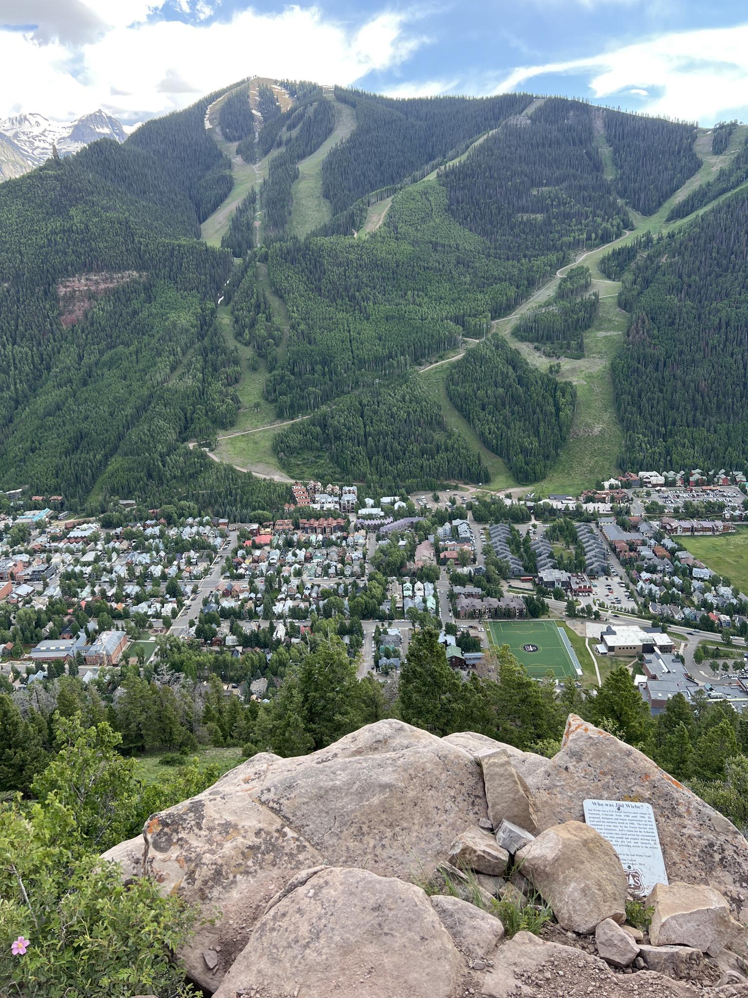 Rocky foreground overlooking a town and a large forested mountain with ski runs.
