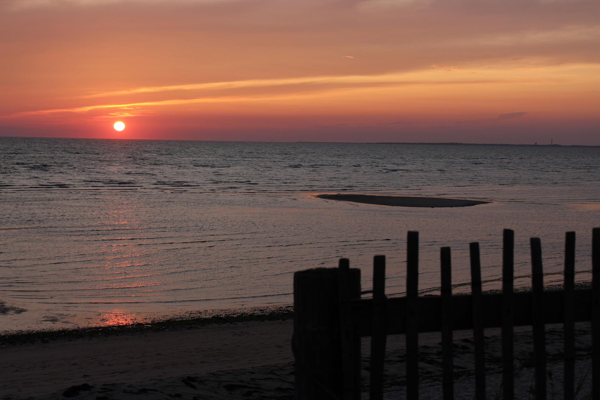 Vibrant sunset over calm water with a sandy beach and wooden fence.