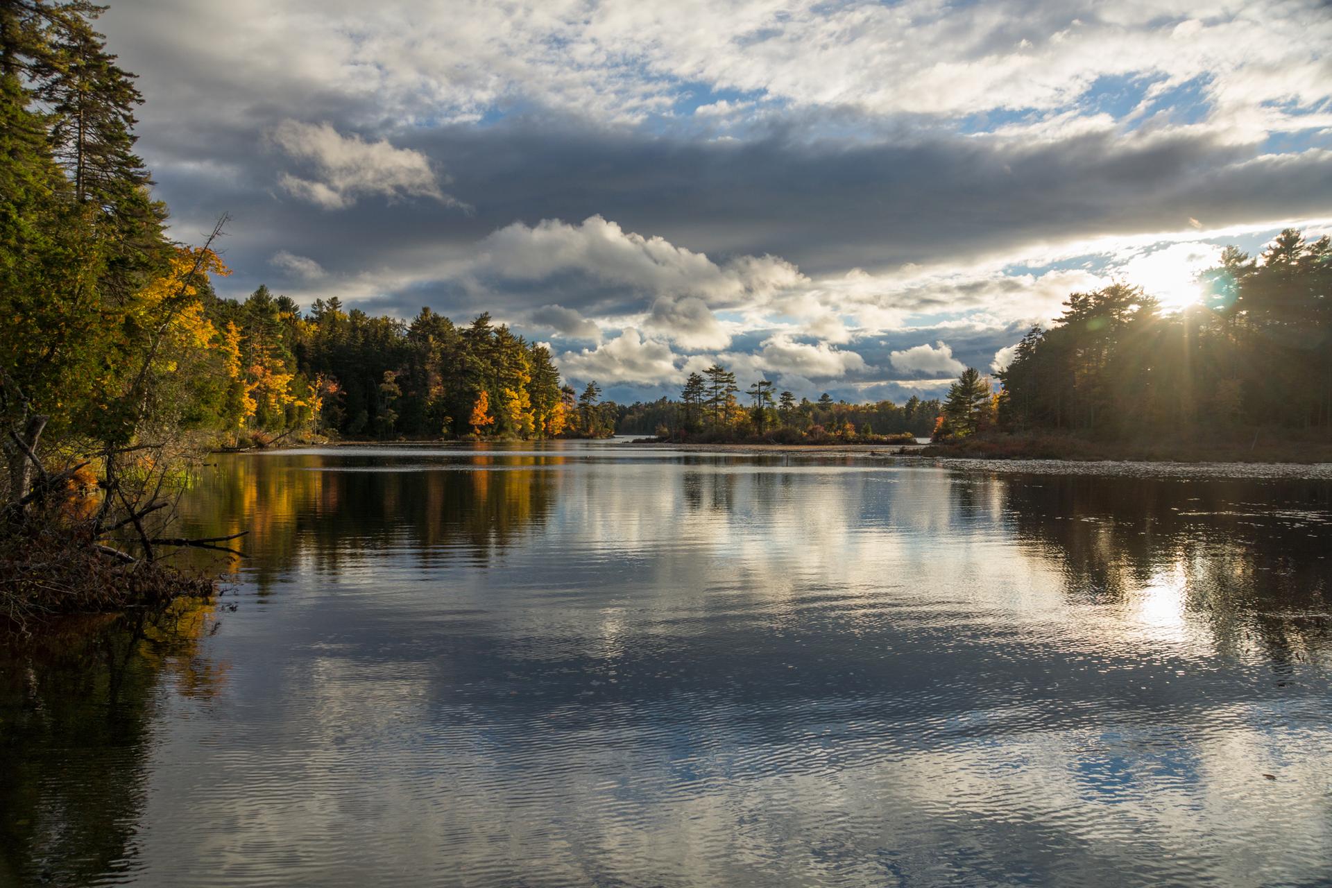Sunlight shines over a calm lake reflecting autumn trees and cloudy sky.