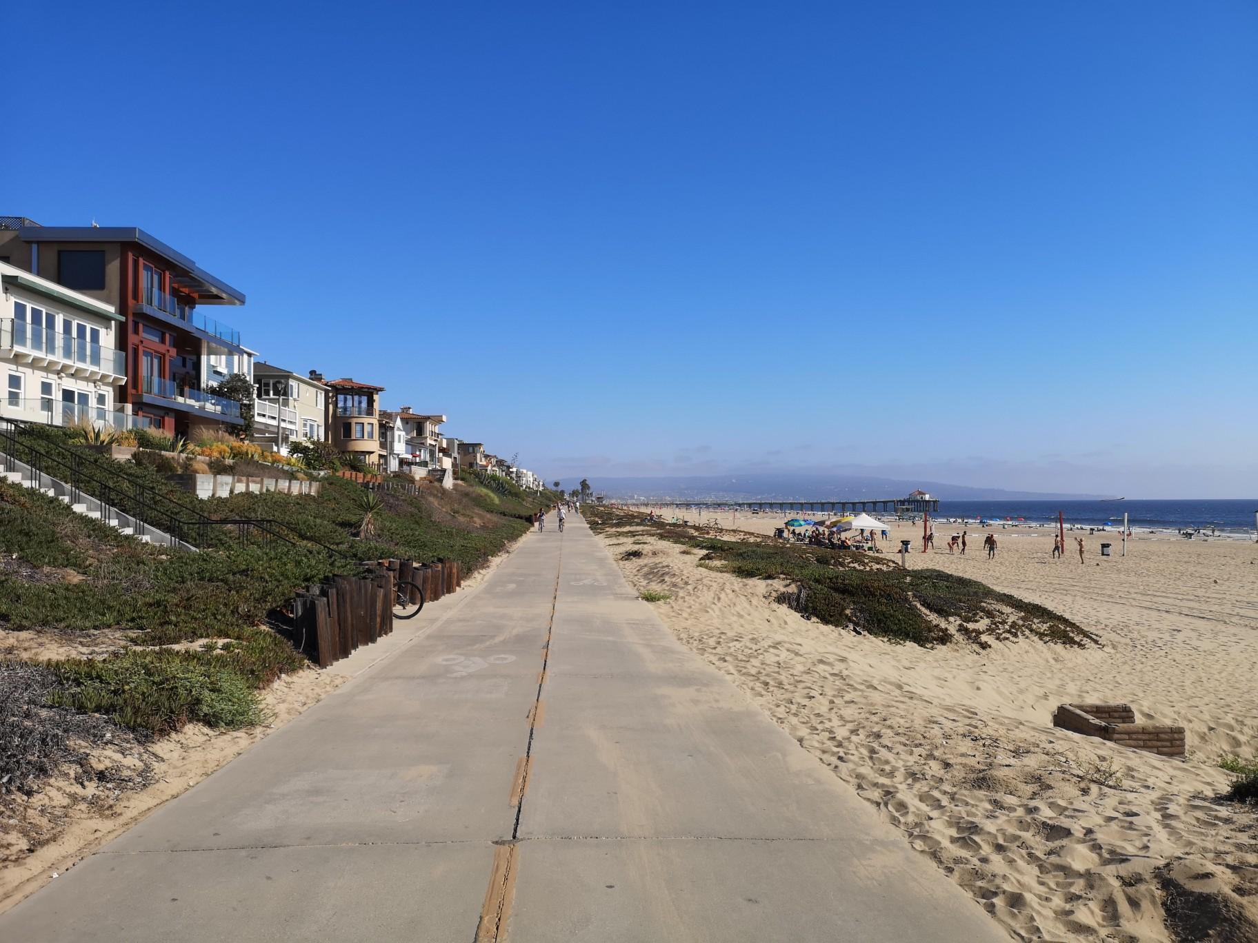Paved path alongside a sunny beach with houses and distant ocean.