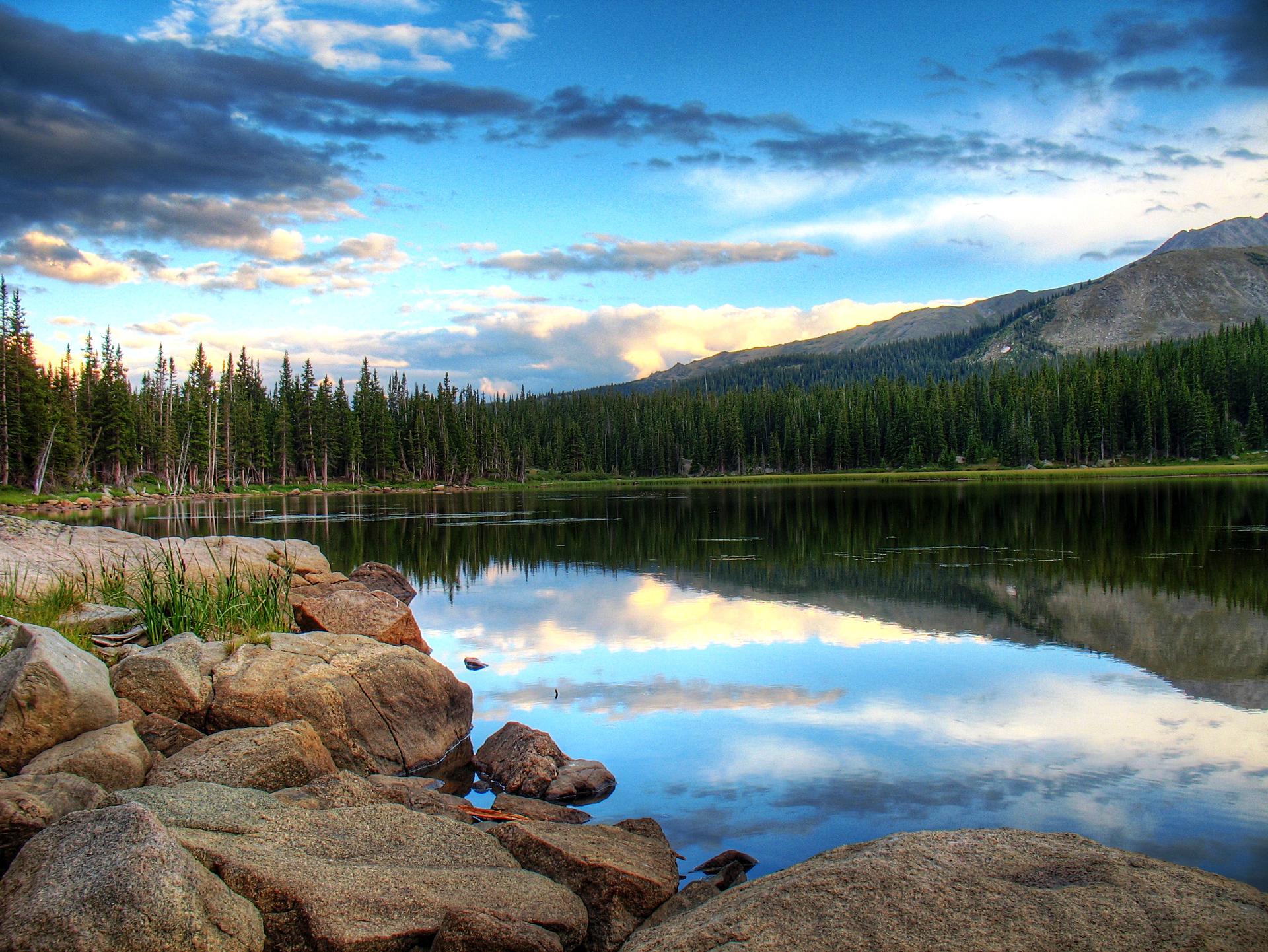 A serene lake reflecting dramatic clouds, with a rocky shore and dense forest.