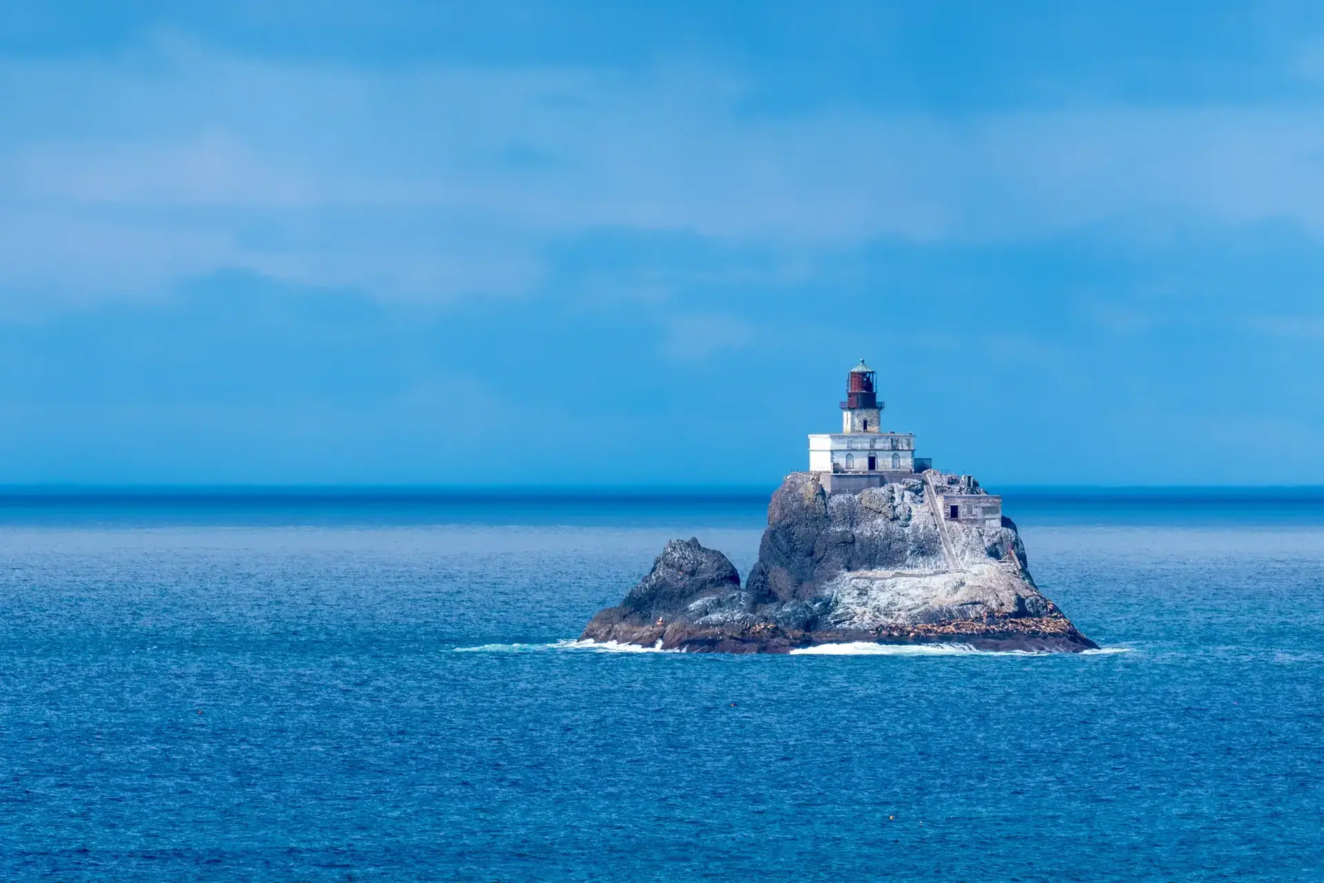 A white lighthouse stands on a rocky island in the vast blue ocean.
