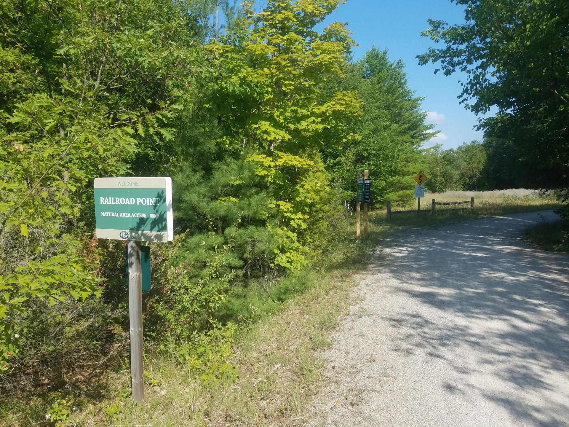 A green sign stands beside a sunny gravel path through green woods.