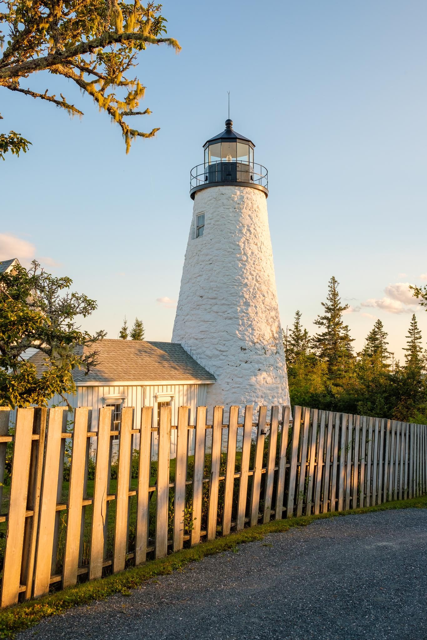 A white lighthouse and building behind a fence in warm, golden hour light. - Blue Hill and Castine Figure-8 from Surry