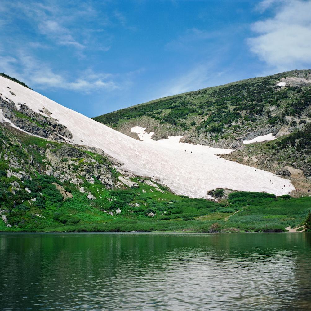 Green lake reflecting a snow-covered mountain slope under a blue sky.