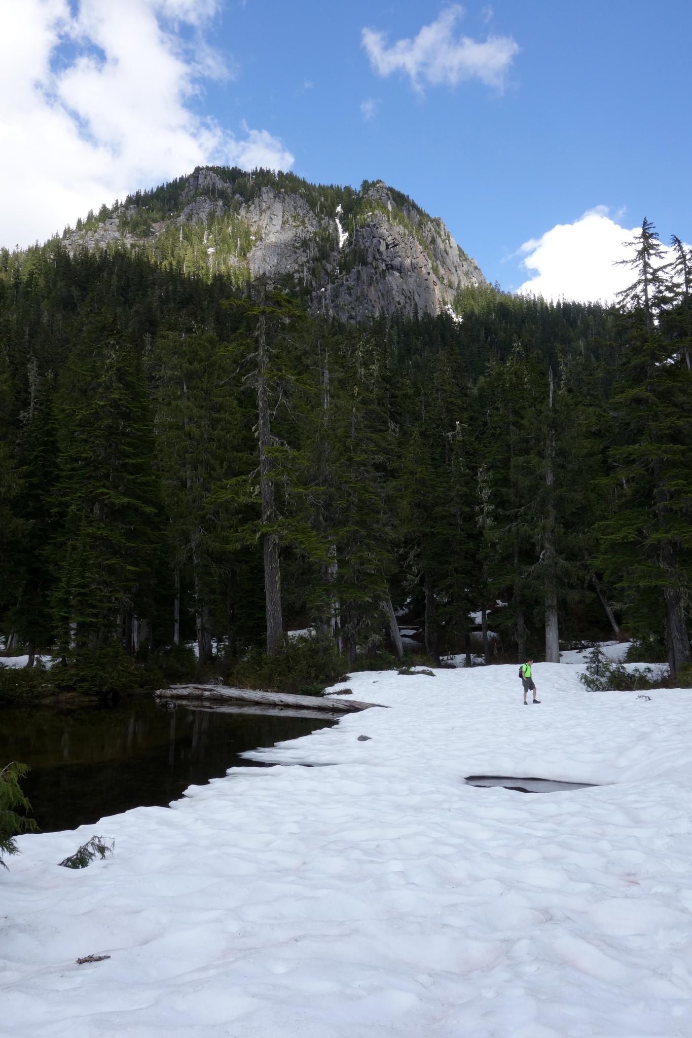 A person stands on snow beside a dark lake, with a forested mountain.