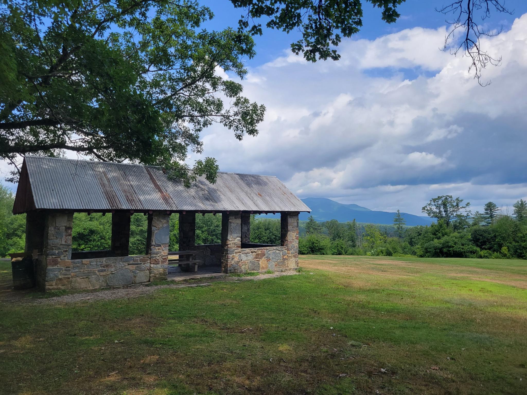 Stone pavilion with metal roof in a grassy field under cloudy sky.
