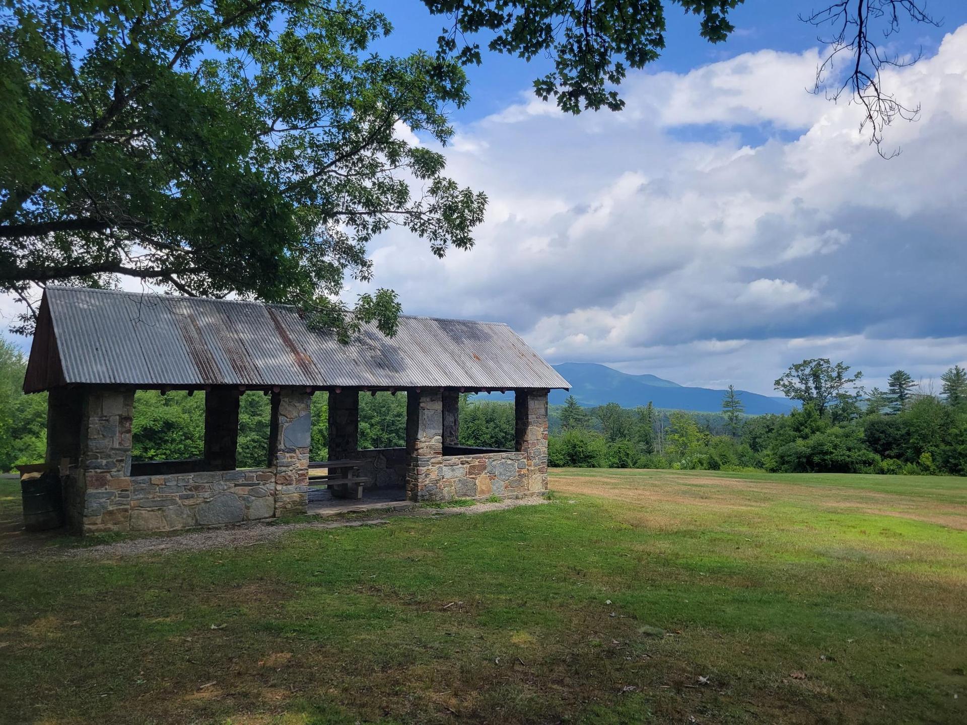 Stone pavilion with metal roof in a grassy field under cloudy sky.