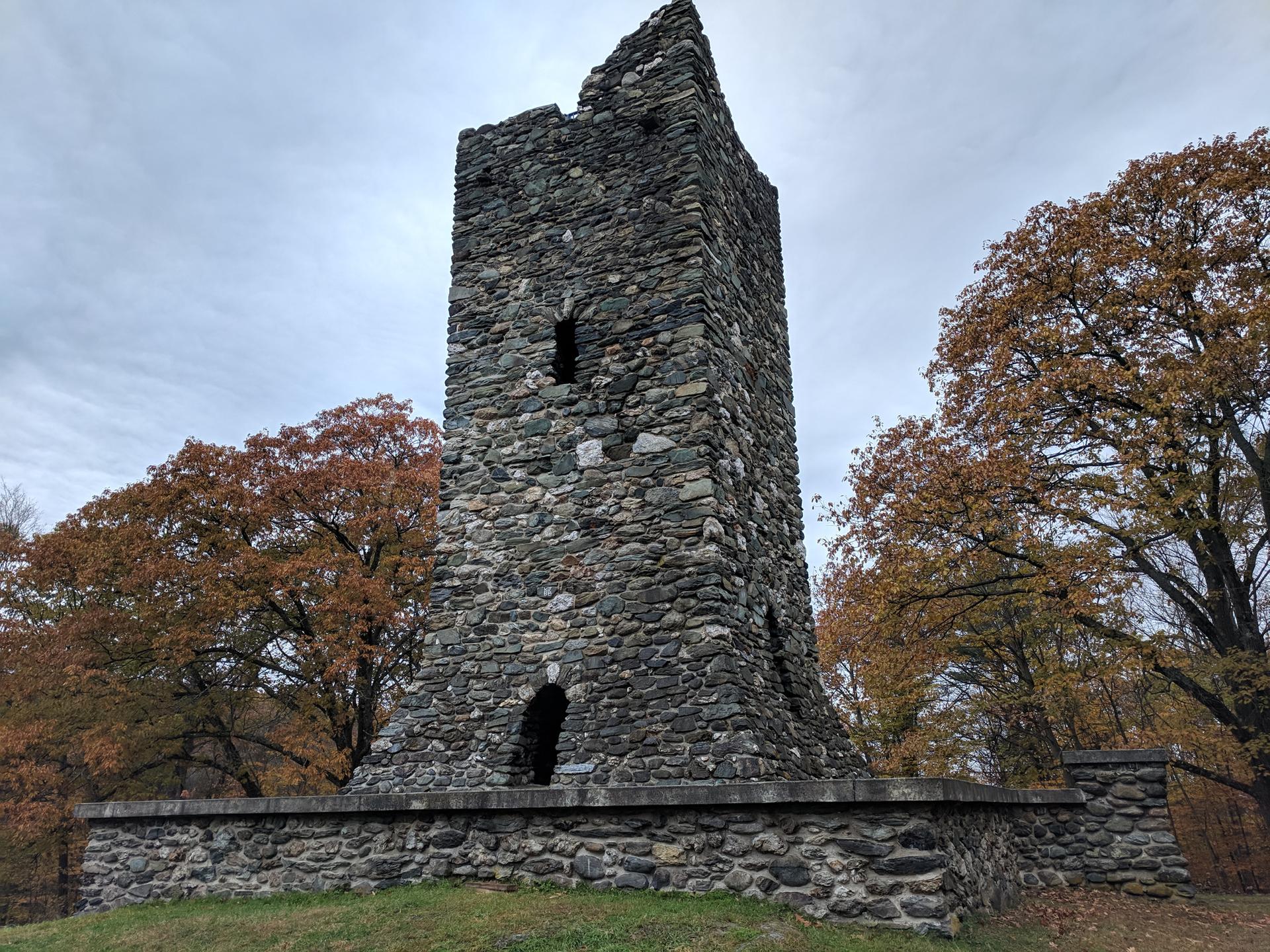 Tall, ruined stone tower surrounded by autumn trees under a cloudy sky.