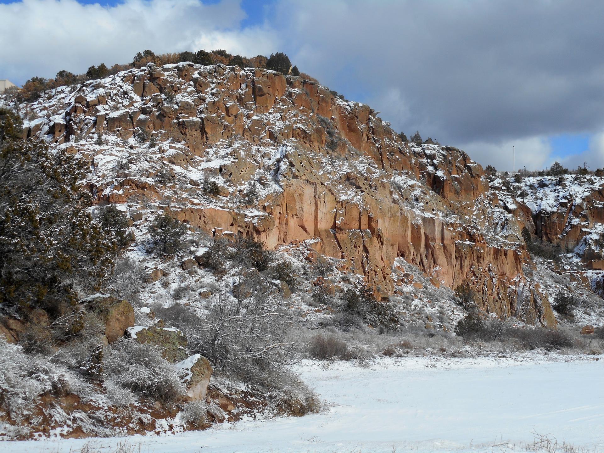 Snow blankets rugged reddish cliffs and a frozen landscape under a cloudy sky.