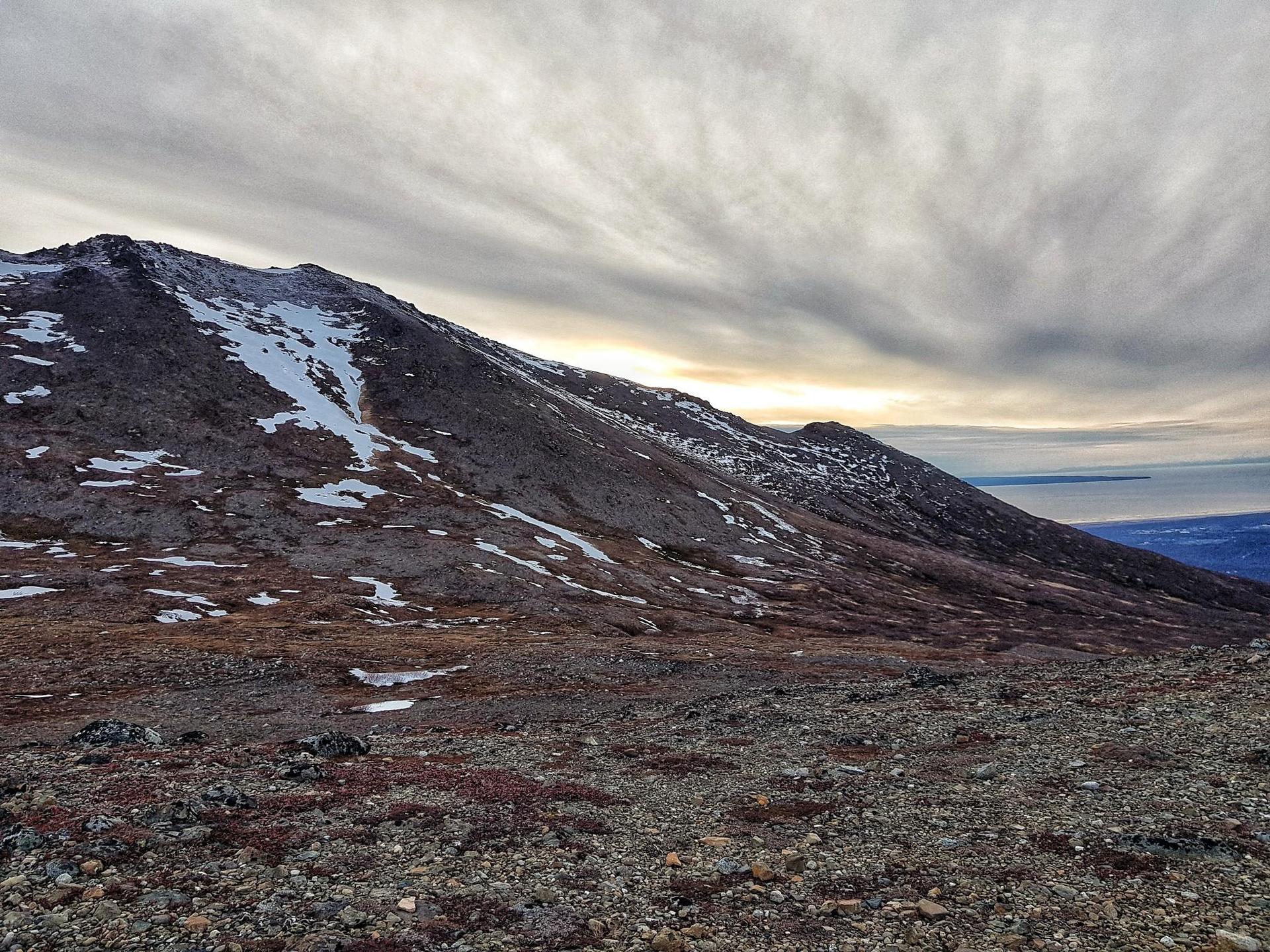 Barren mountain landscape with snow, cloudy sky, and distant water at sunset.