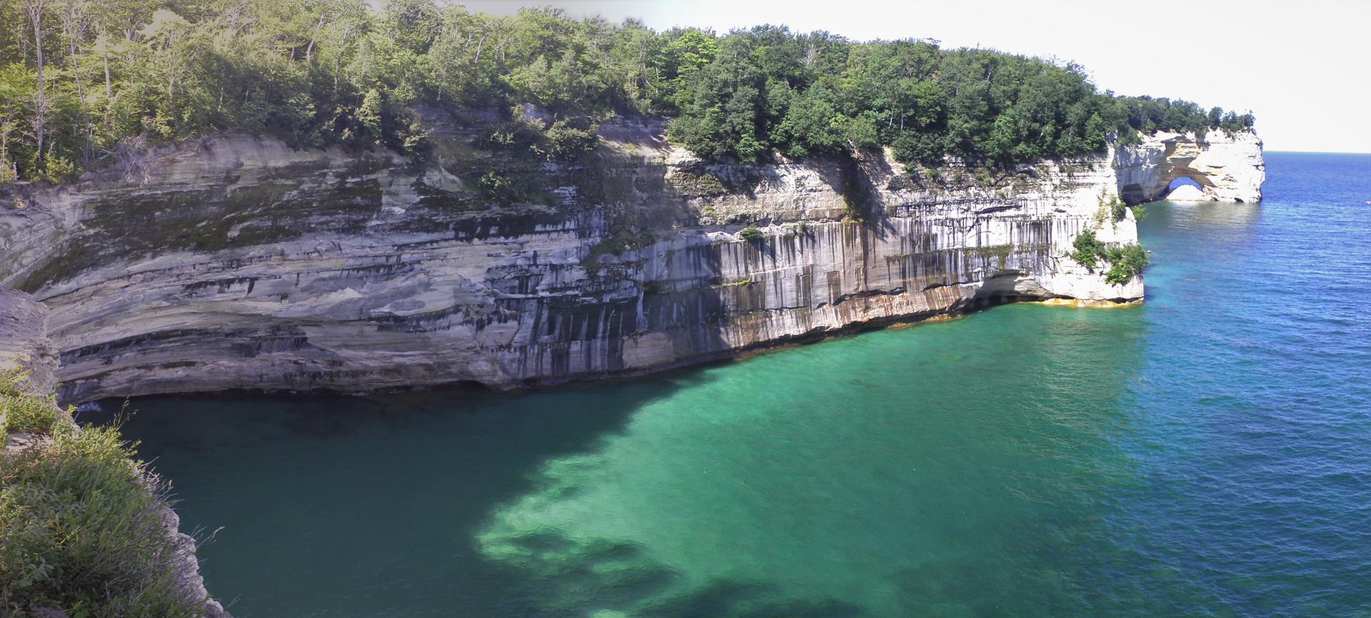Panoramic view of a forested cliff over turquoise water with rock formations.