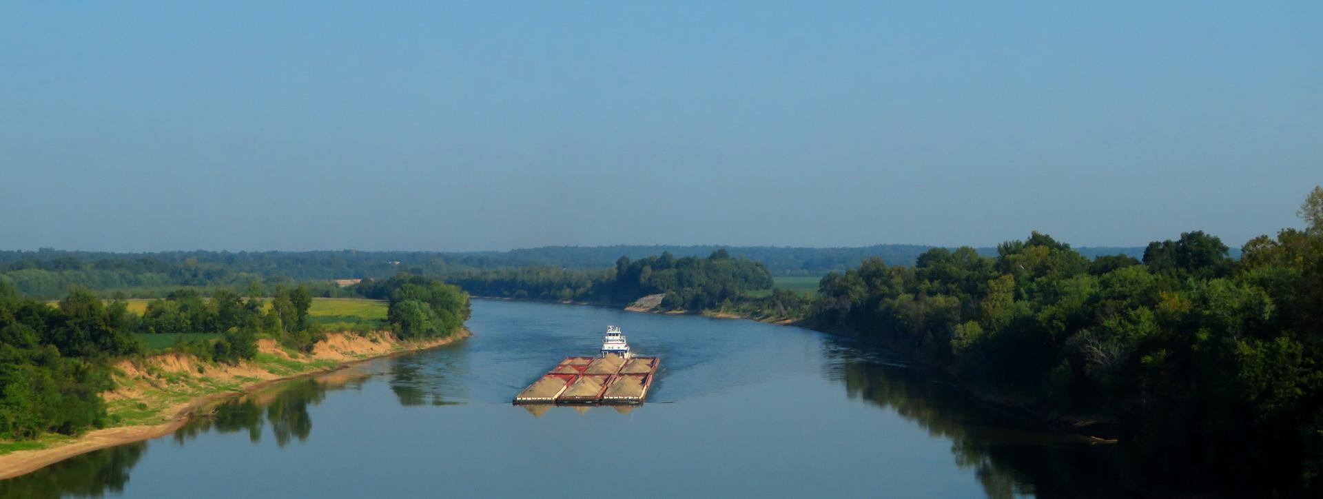 A large barge floats down a wide river with lush green banks.