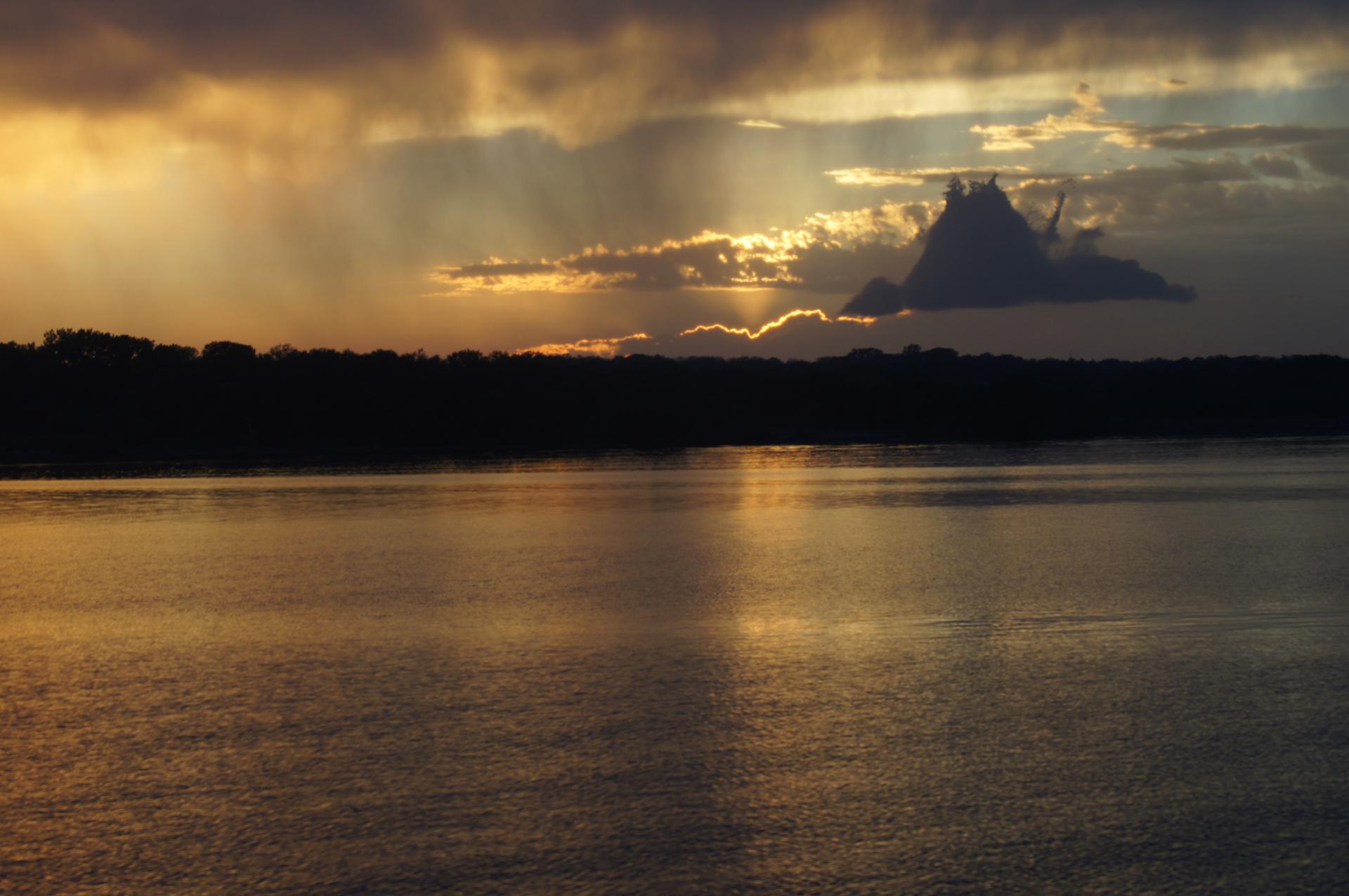Golden light breaks through dark clouds over a calm lake and distant treeline.