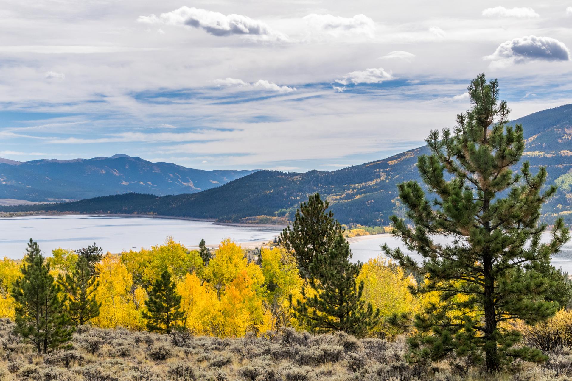 Scenic lake and mountains with vibrant yellow and green trees under a cloudy sky.