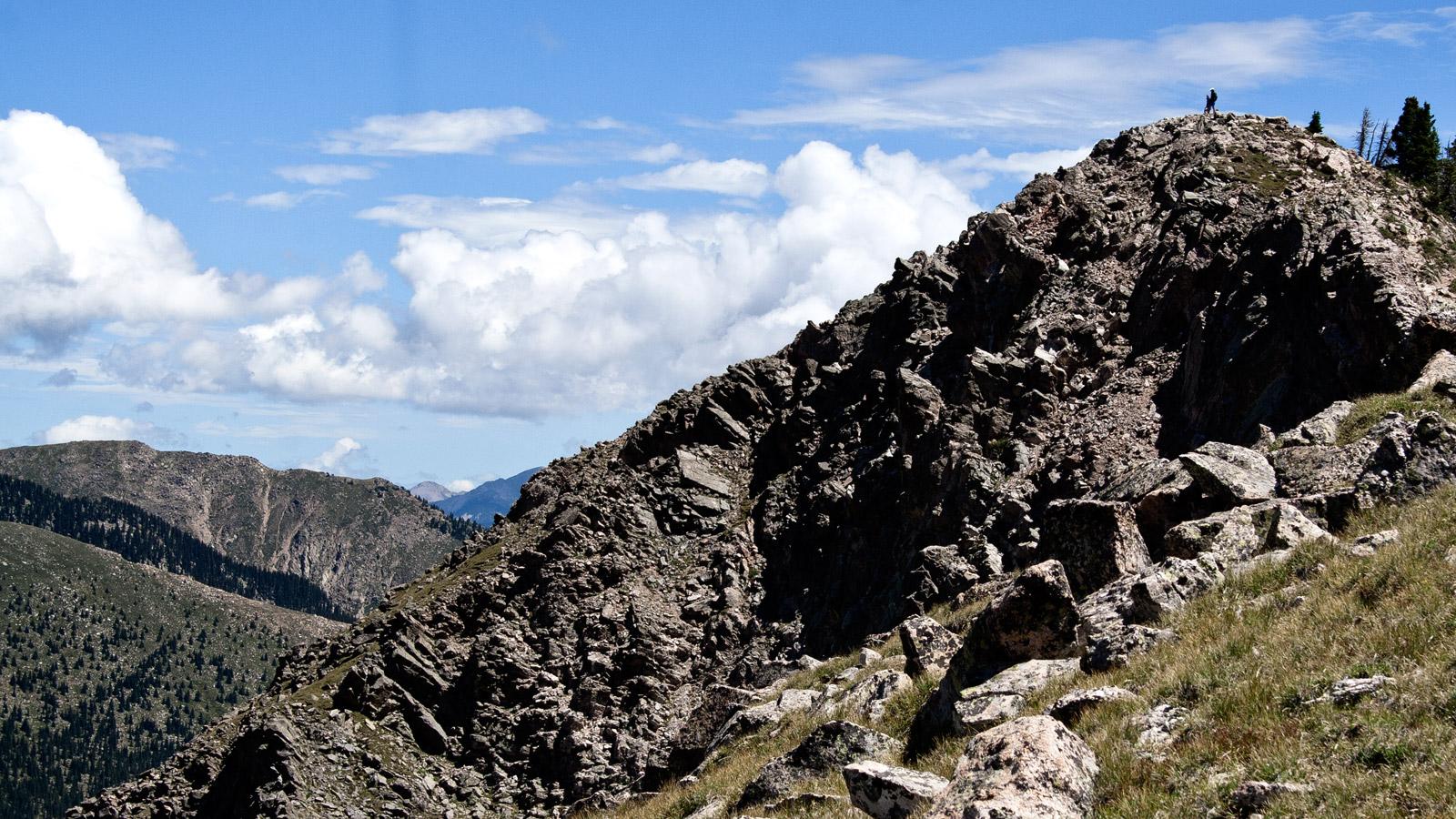 A person stands atop a rugged, rocky mountain peak under a cloudy sky.