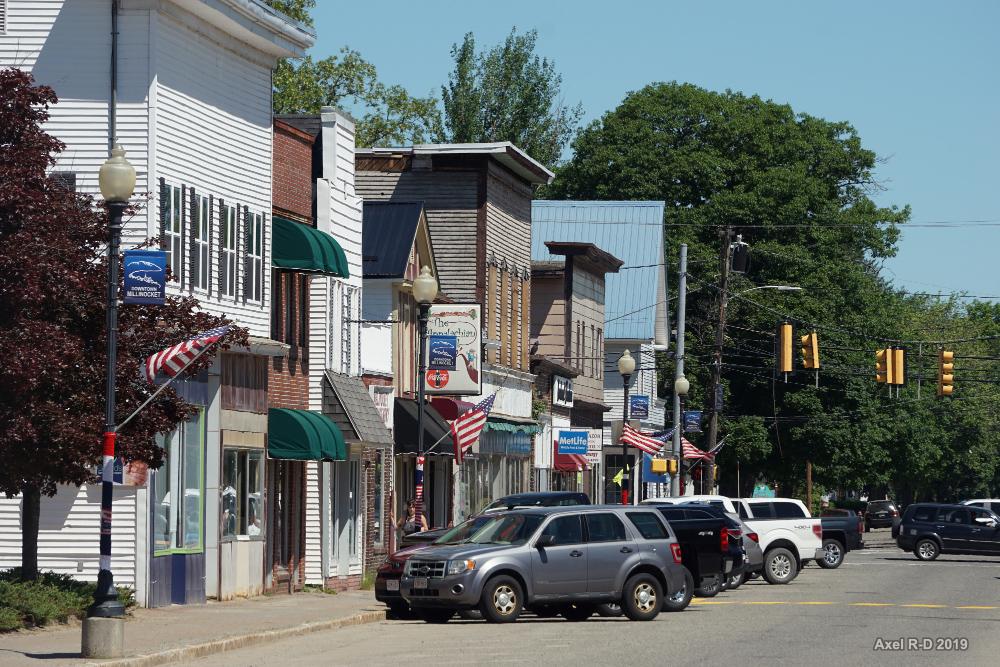 Bright daylight illuminates a street lined with shops, flags, and parked vehicles.