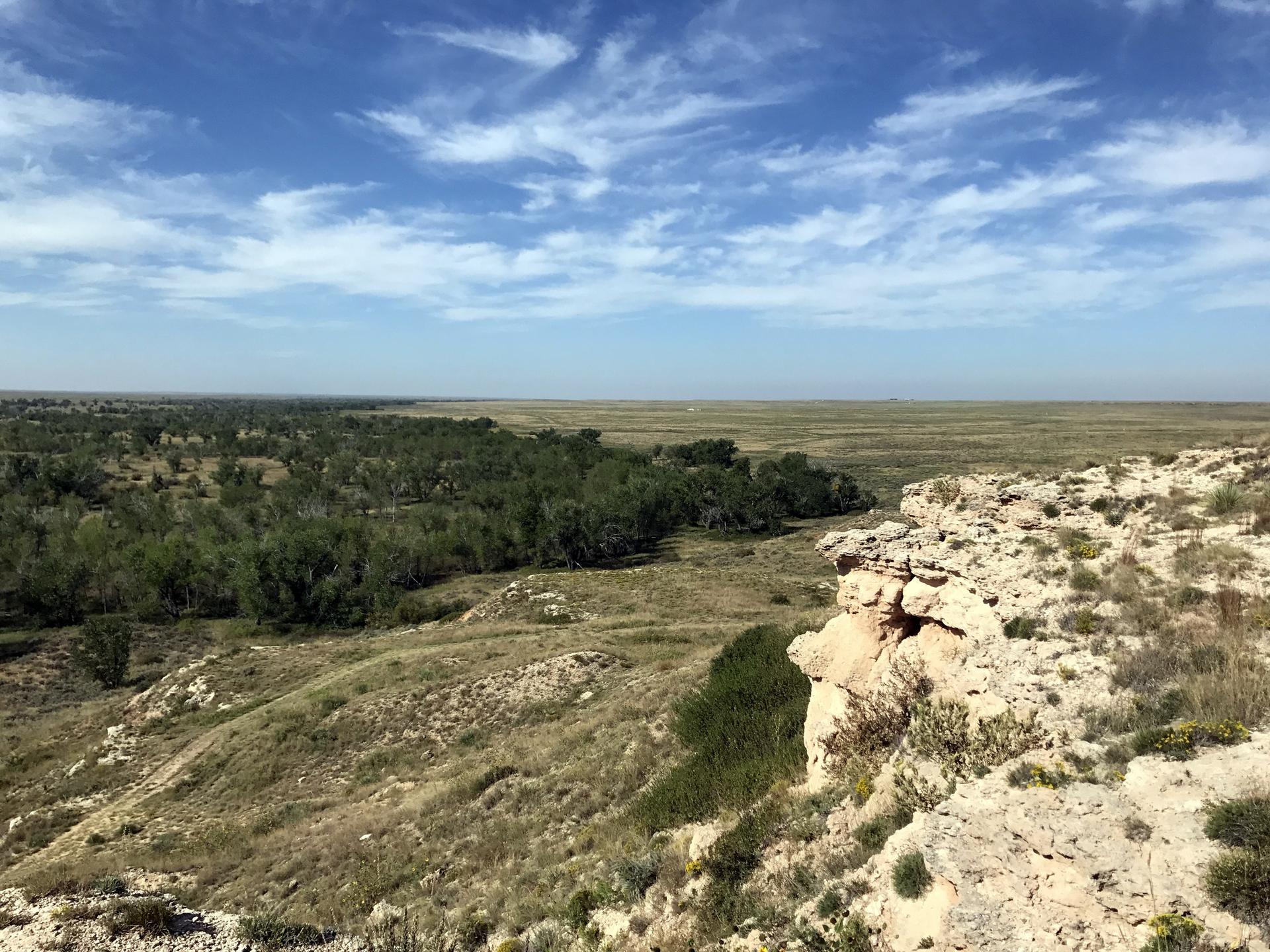 Expansive view of a green valley and dry plains from a rocky cliff under a blue sky.
