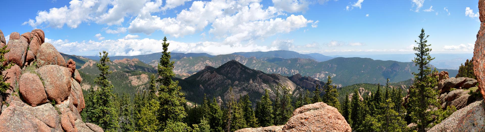 Panoramic mountain view with evergreen trees, rocky foreground, and a bright, cloudy sky.