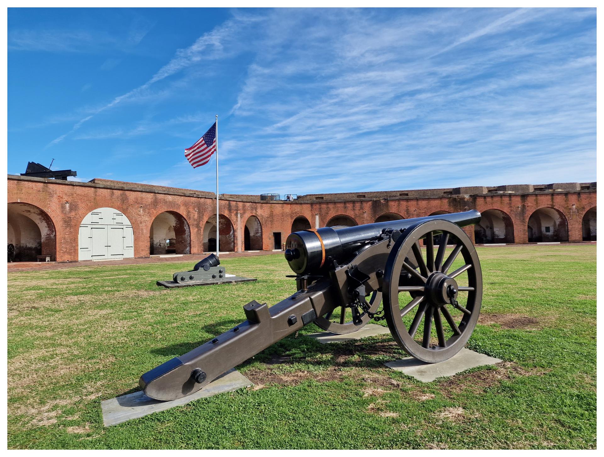Large cannon on green grass with a fort and flag under a blue sky.