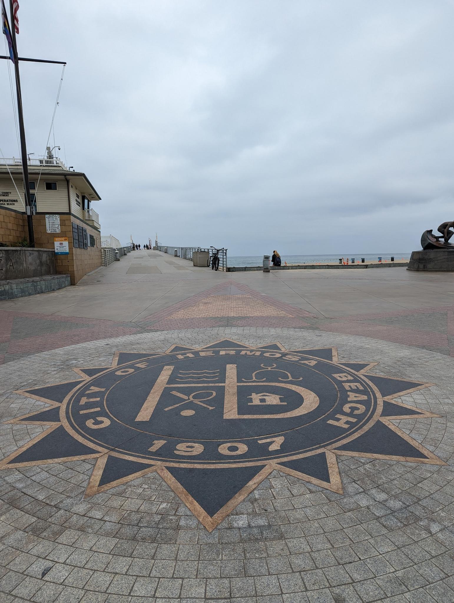Paved promenade with a large emblem leading to a pier under cloudy skies.