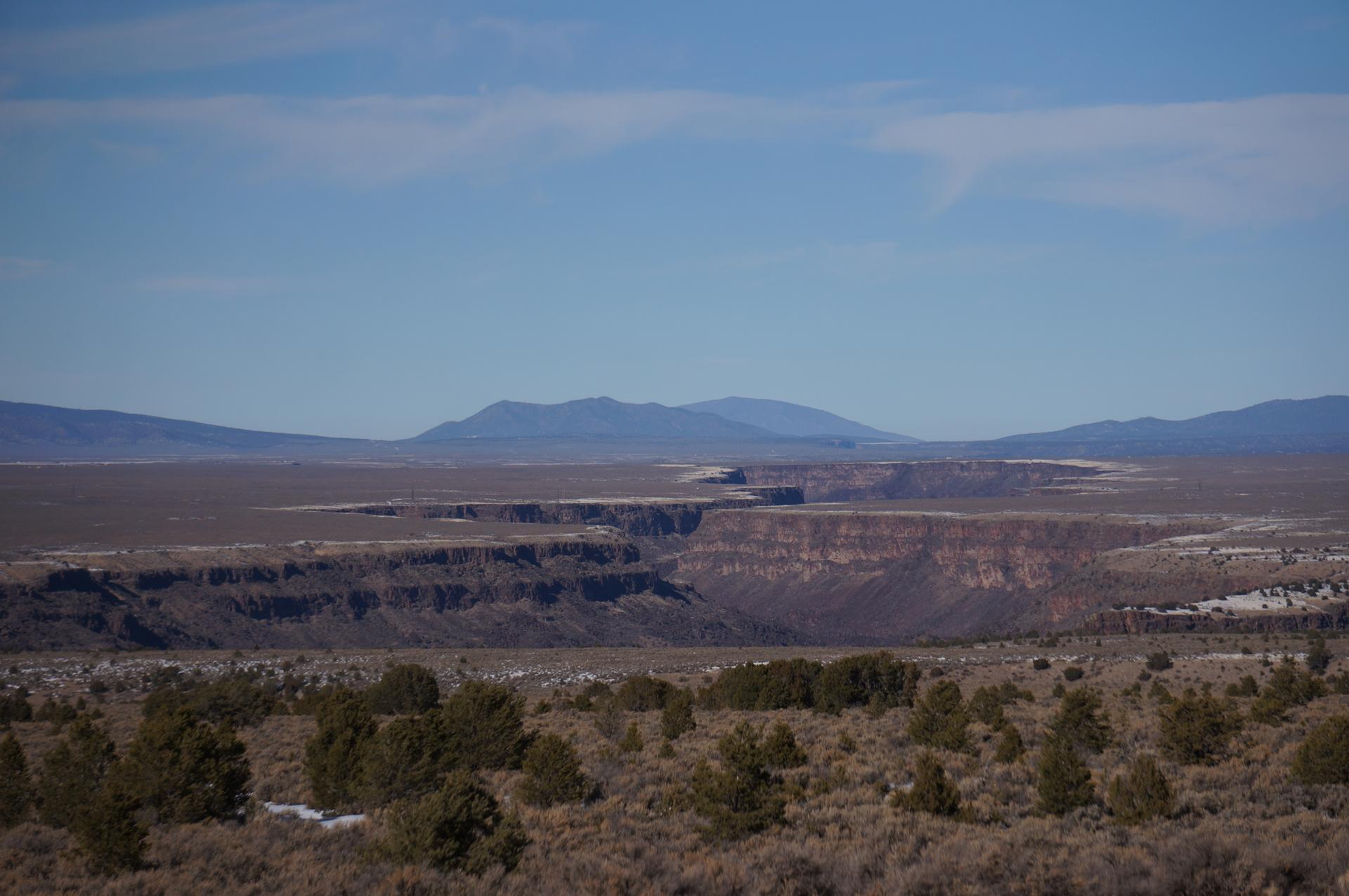 Vast desert canyon with layered rock formations and distant mountains under a hazy sky.