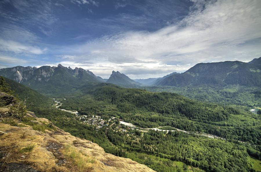 Panoramic view of a vast forested mountain valley with a river and dramatic sky.