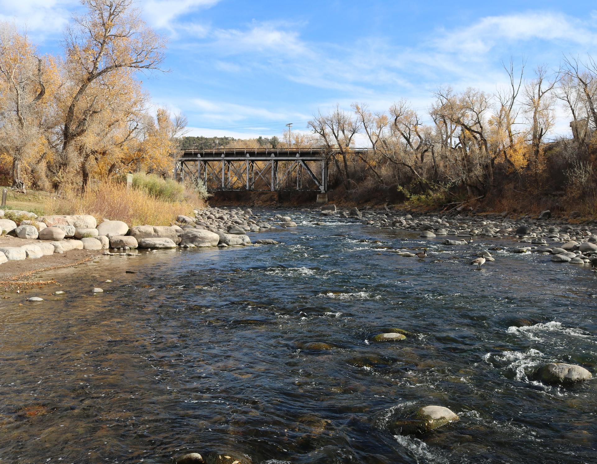 A rocky river flows under a bridge with bare trees on a sunny day.