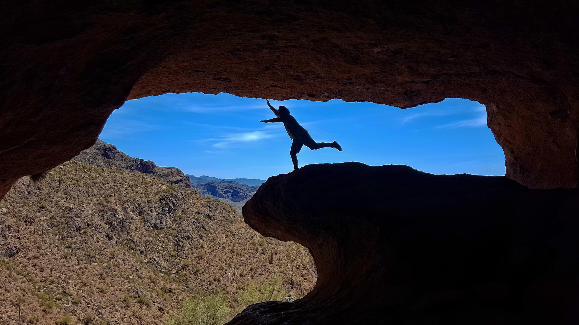 Silhouetted person balancing on a cave ledge against a bright blue sky and mountains.