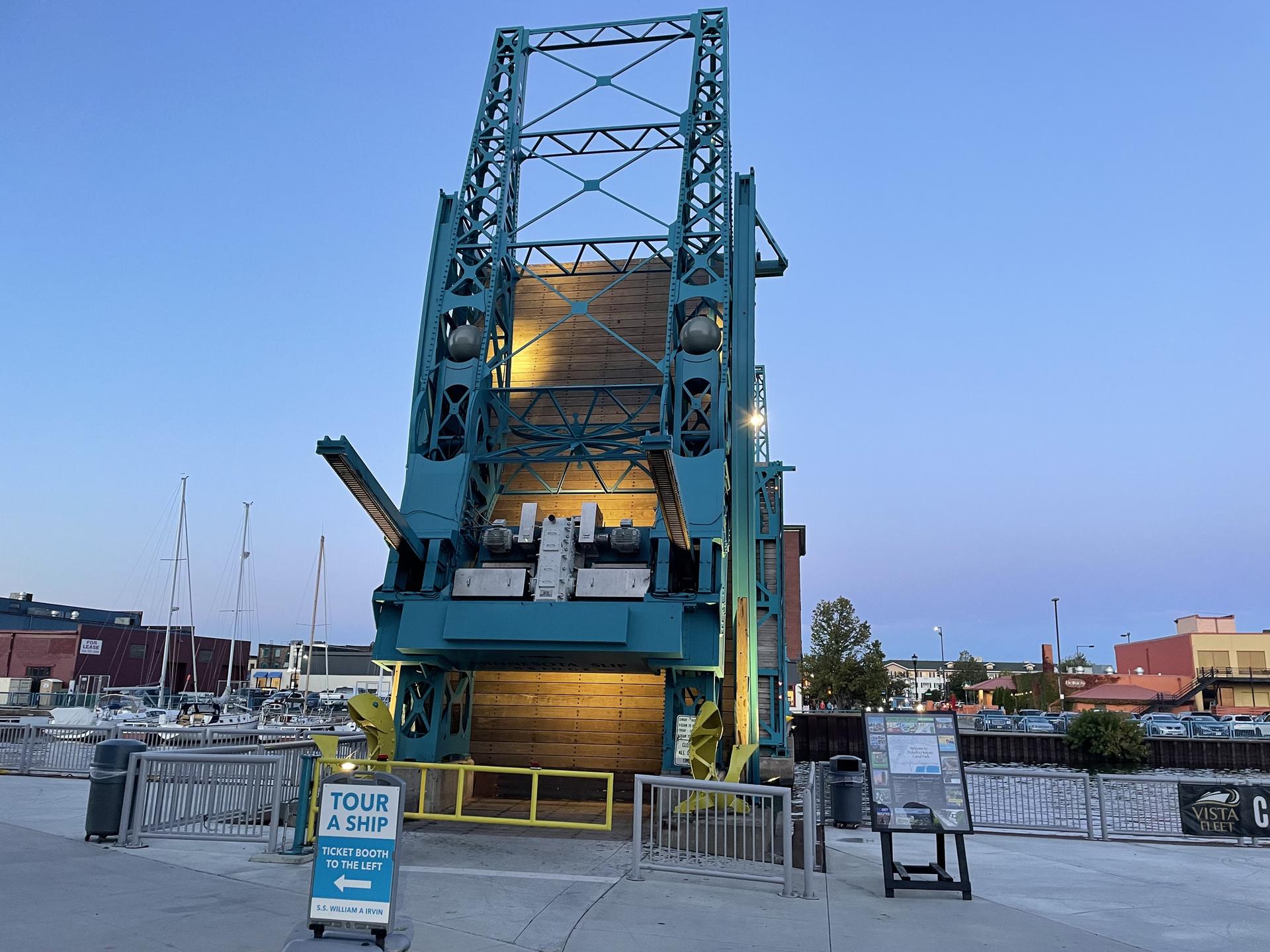 A large teal lift bridge structure at dusk, with boats in the background.