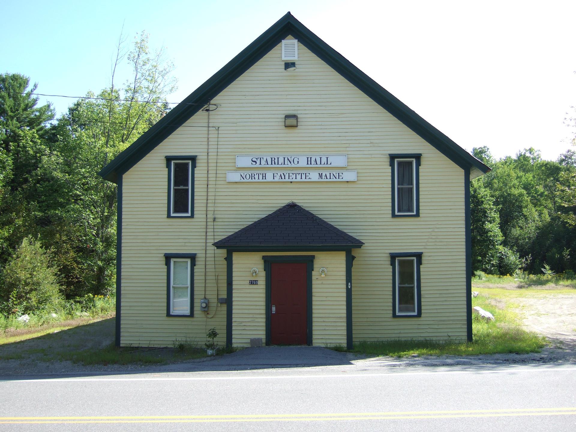 A light yellow building with a dark roof and red door on a sunny day.