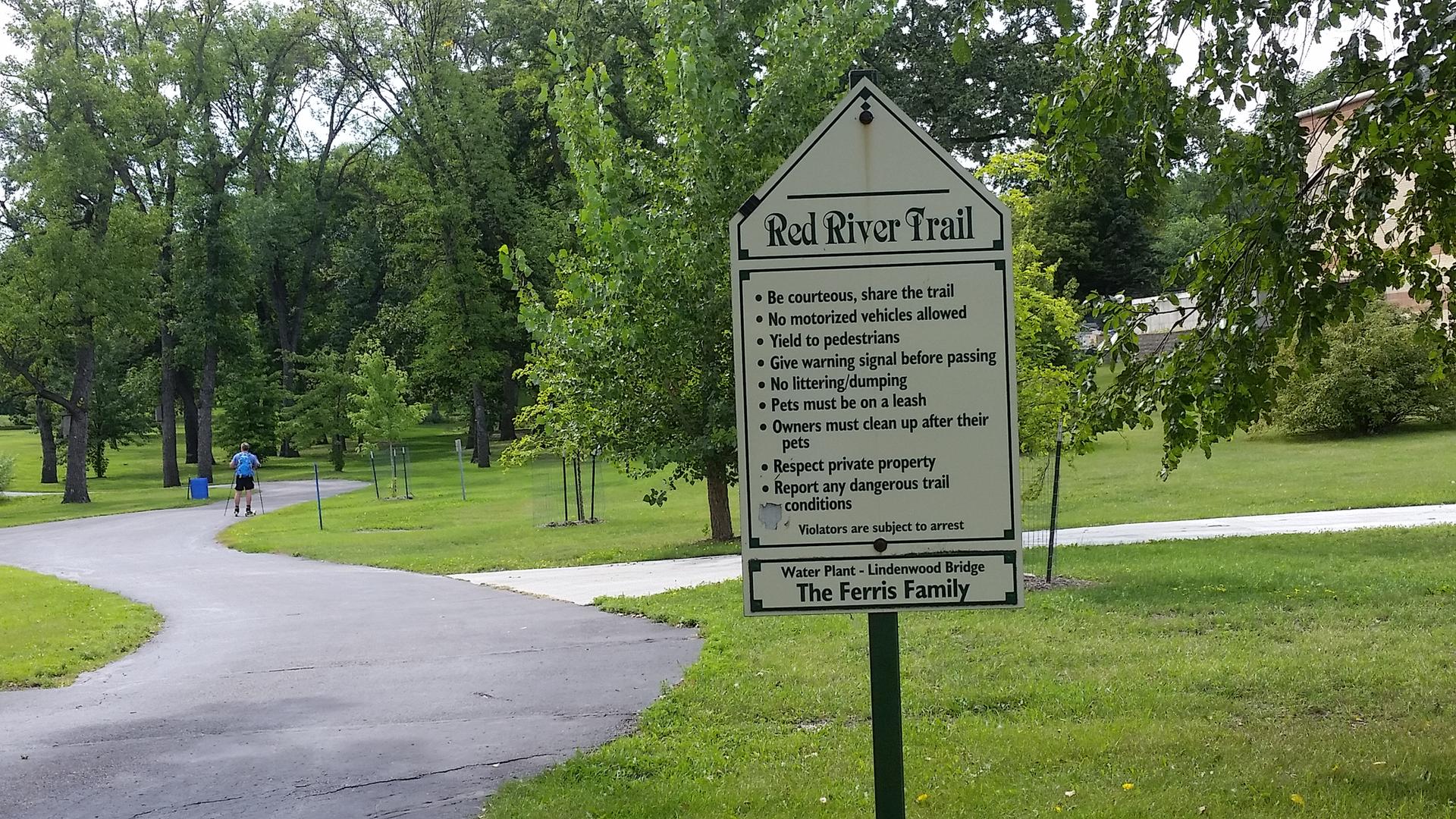 Trail sign beside a paved path with a person walking in a wooded park.