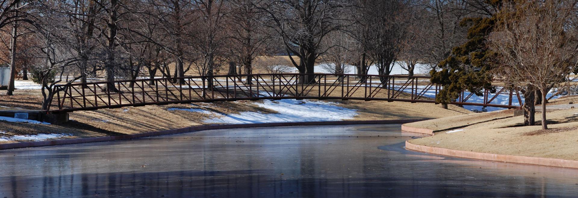 A curved wooden bridge spans still water with bare trees and snow.