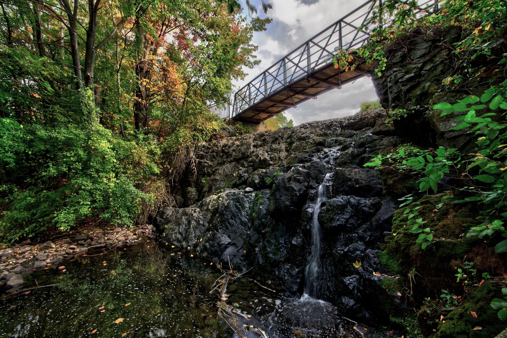 A waterfall cascades over dark rocks under a metal bridge, surrounded by lush trees.