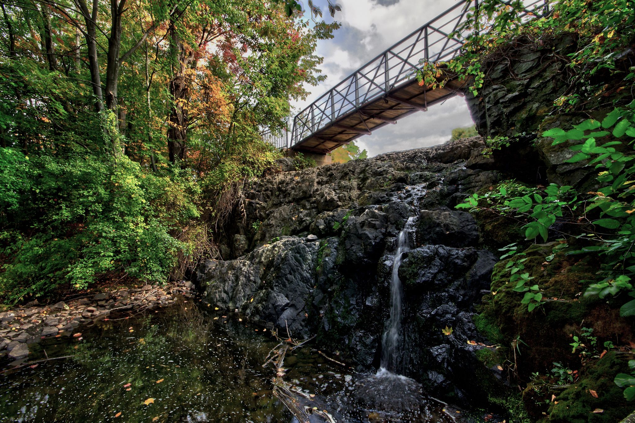 A waterfall cascades over dark rocks under a metal bridge, surrounded by lush trees. - CTfastrak and Mill Pond Trail Loop
