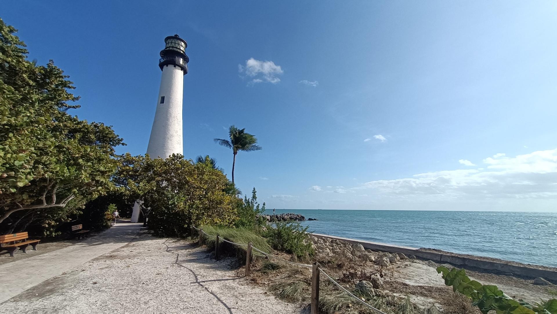 A tall white lighthouse stands beside a path and the blue ocean.