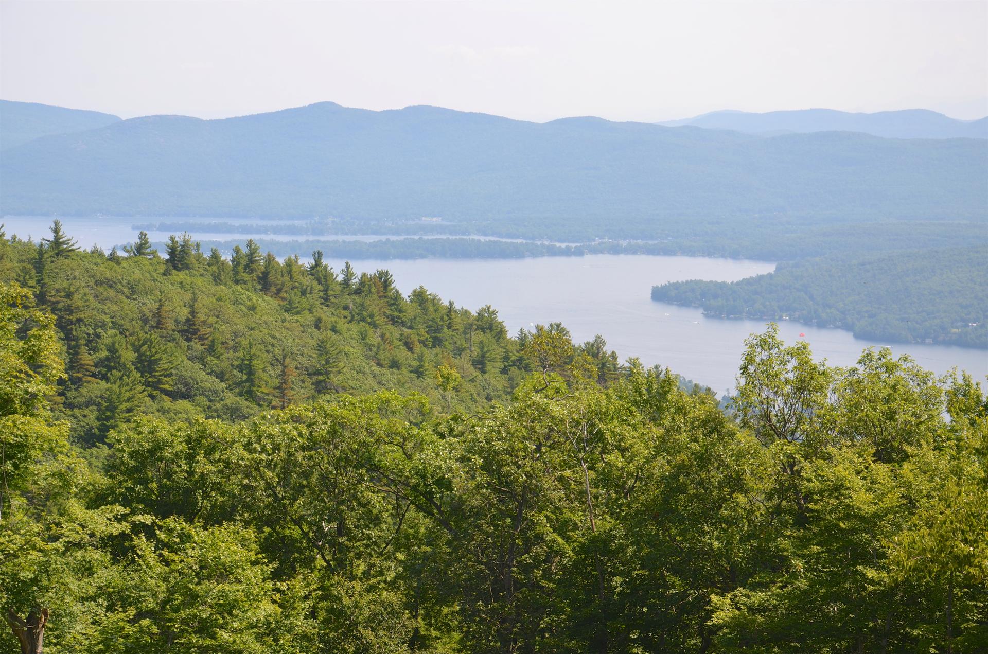 Panoramic view of a lake surrounded by green forests and distant hazy mountains.