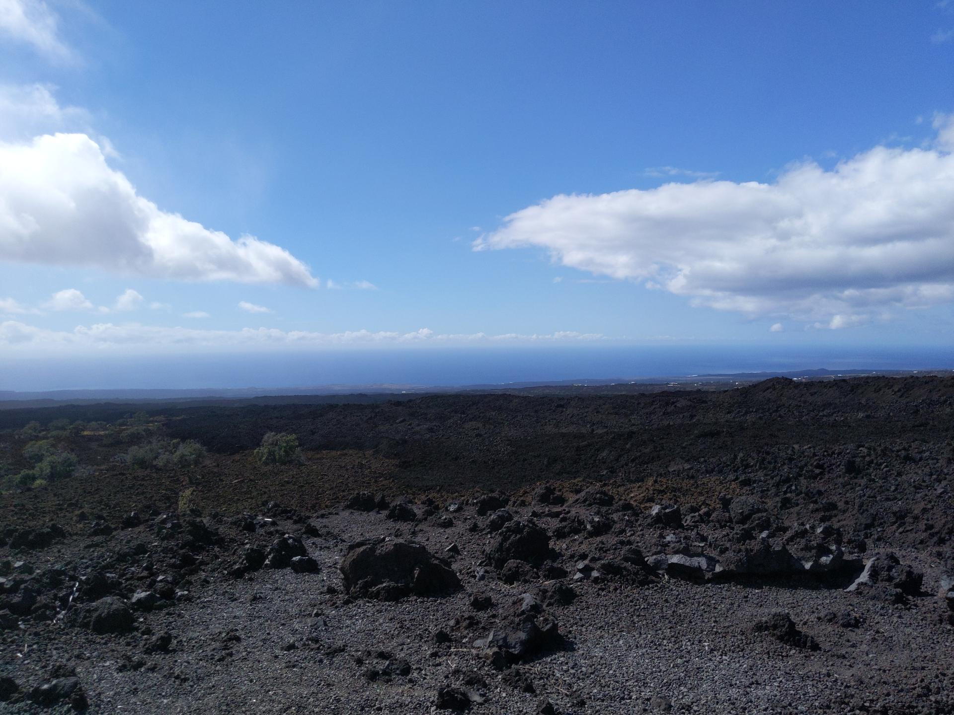 Vast dark rocky landscape under a bright blue sky with scattered clouds, distant ocean.