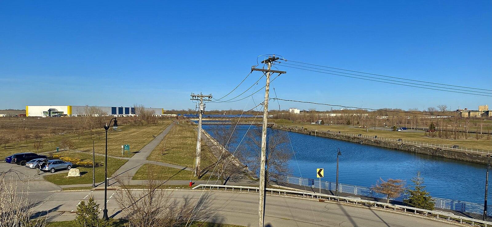 Wide canal with industrial buildings, power lines, and parked cars.