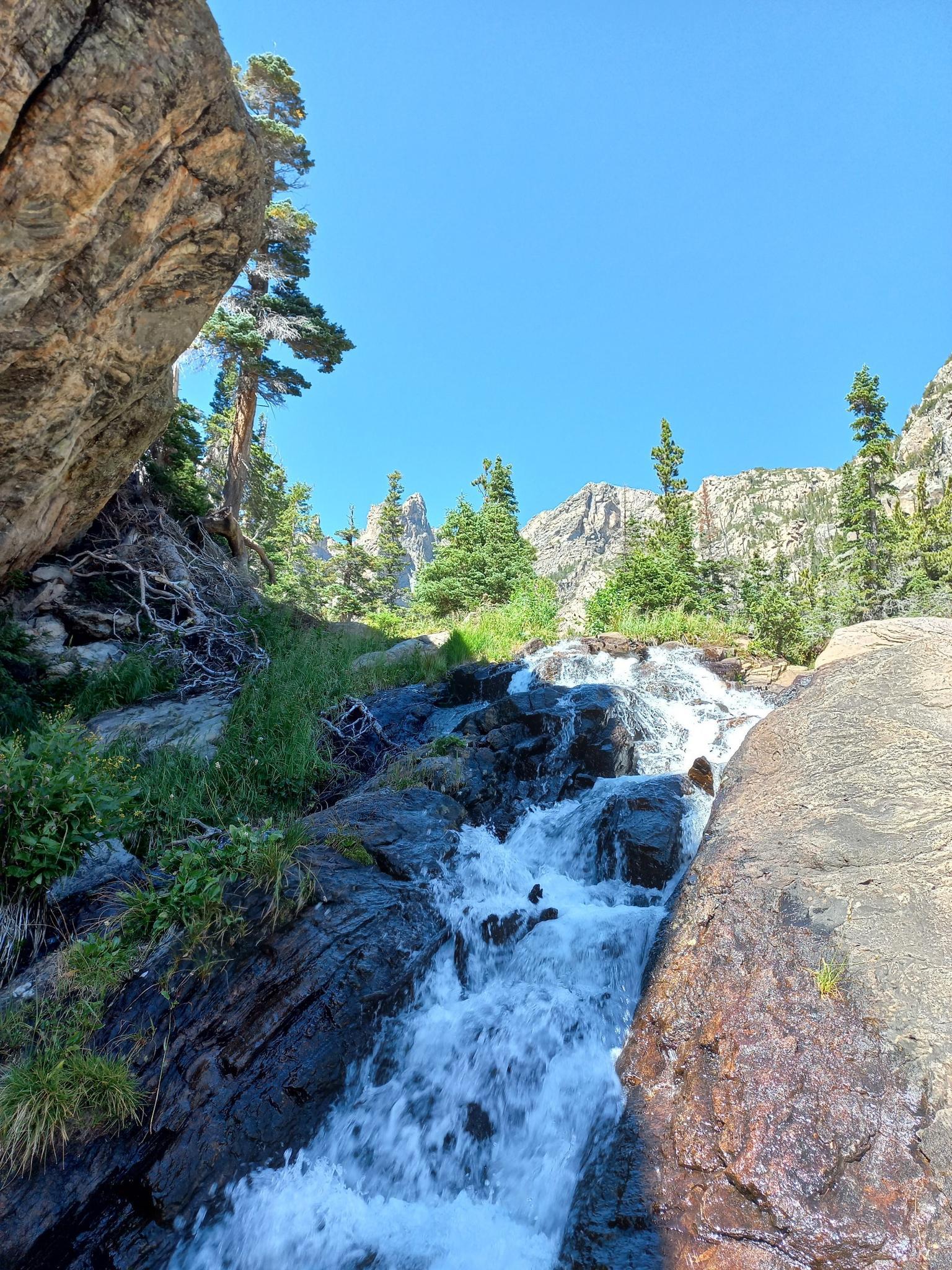 Small waterfall cascading over rocks in a sunny, rugged mountain landscape.