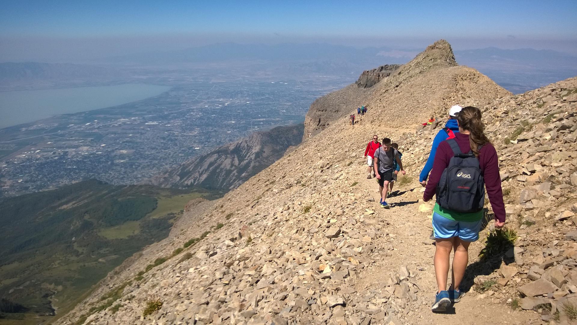 Hikers on a rocky mountain trail overlooking a distant lake and city.